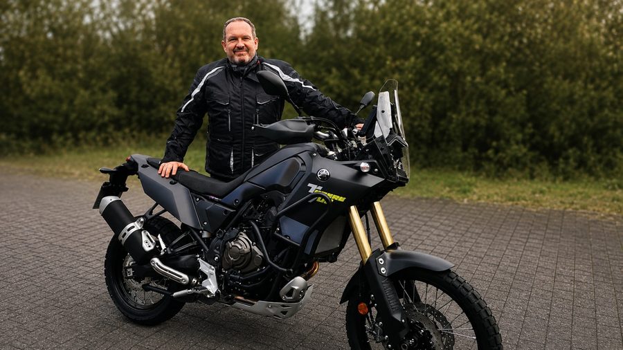 Claus Schertz steht neben seinem Motorrad, im Hintergrund grüne Hecken